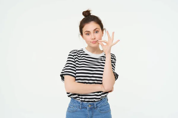 Portrait of brunette woman seals her mouth with promise not to tell anyone, zipping gesture, standing over white background.