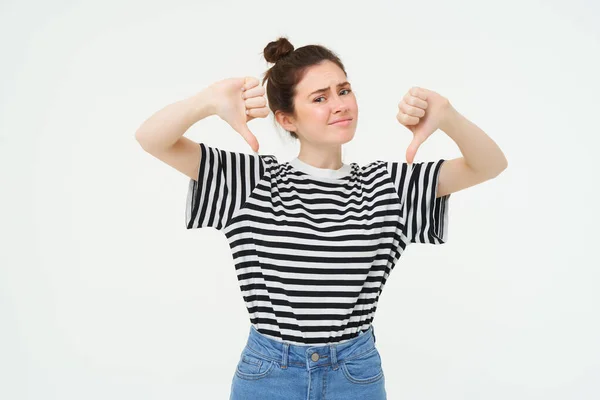 Young woman gives negative feedback, shows thumbs down, disapproves product, dislikes smth bad, stands over white background.