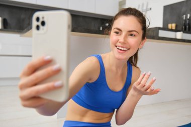 Genç fitness kızı portresi, blogcu evden antrenmanını kaydediyor, akıllı telefonda selfie için poz veriyor, kamerada yoga yapıyor, mavi spor sutyen ve taytla oturuyor..