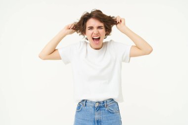 Woman screams, pulls out her hair and shouts, stands frustrated, angry and pissed-off, stands over white studio background.