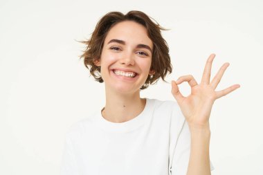 Tatmin olmuş, gülümseyen genç bir kadının yakın portresi, tamam, tamam bir jest gösterir, bir şey tavsiye eder, olumlu geri bildirim verir, evet der, beyaz arka plan.