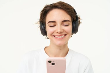 Mutlu, gülümseyen güzel bir kadının portresi, kablosuz kulaklıkla müzik dinlemek, akıllı telefona bakmak, canlı uygulamadan şarkı seçmek, cep telefonu tutmak, beyaz arka planda durmak..