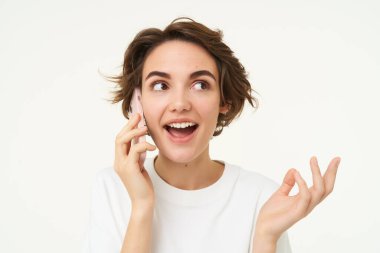 Mutlu, konuşkan genç bir kadının portresi cep telefonuyla konuşuyor, akıllı telefon kullanıyor, birini arıyor, beyaz stüdyonun arka planında dikiliyor..