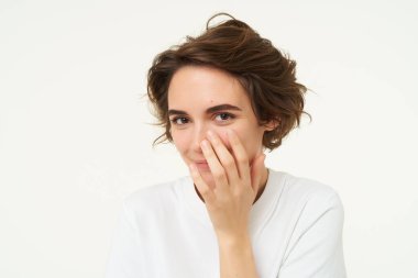 Şirin, genç, esmer bir kadına yakın, yüzünü örtüyor, gülüyor ve gülümsüyor, utangaç ve aptal görünüyor, kıkırdıyor, beyaz arka planda duruyor..
