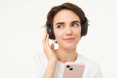Samimi genç bir kadının portresi, kablosuz kulaklıkla müzik dinliyor, akıllı telefon tutuyor, cep telefonuyla video izliyor, beyaz arka planda duruyor..