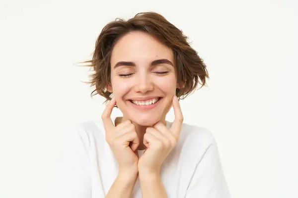 stock image Portrait of brunette woman smiling, shuts her eyes and touches dimples on her cheeks, stands over white background. Copy space