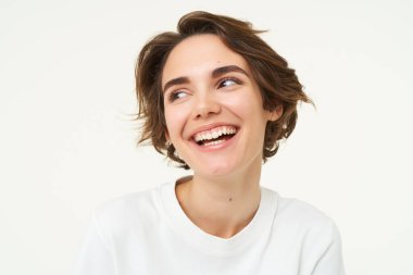 Esmer kadının yakın portresi gülüyor ve gülümsüyor, gerçek duygularını ifade ediyor, beyaz stüdyo arka planına karşı poz veriyor..