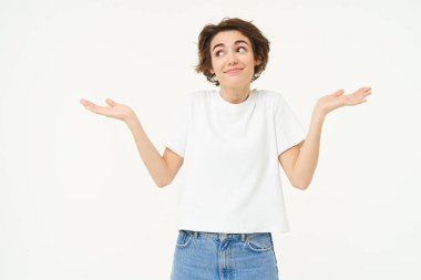 Kısa saçlı bir kızın portresi, omuz silkiyor, habersiz görünüyor, ne yapacağını bilmiyor, beyaz stüdyo arka planında duruyor..
