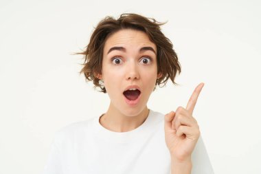 Heyecanlı kızın bir fikri var, çözümü düşünüyor, bir parmağını gösterip gülümsüyor, beyaz arka planda duruyor..