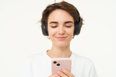 Samimi genç bir kadının portresi, kablosuz kulaklıkla müzik dinliyor, akıllı telefon tutuyor, cep telefonuyla video izliyor, beyaz arka planda duruyor..