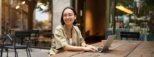 stock image Portrait of asian girl works in cafe, uses laptop and sits outdoors on street. Digital nomad and online learning concept.