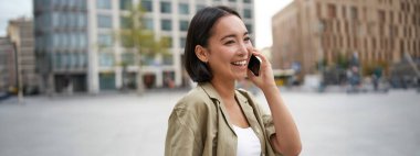 Modern Asyalı genç kız cep telefonuyla konuşuyor, şehir caddesinde telefon kullanıyor. Akıllı telefondan birini ararken gülümseyen bir kadın..