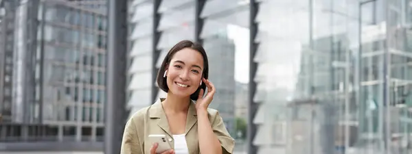 Mladá Usměvavá Asijská Žena Bezdrátových Sluchátkách Chodí Ulici Chytrým Telefonem — Stock fotografie