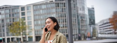 Gülümseyen Asyalı bir kızın dikey görüntüsü, telefonla konuşması ve sokakta yürümesi. Cep telefonuyla konuşan genç bir kadın..