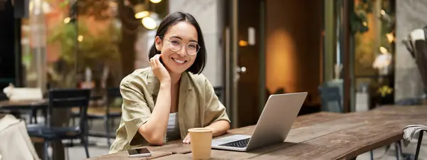 stock image Portrait of smiling girl in glasses, sitting with laptop in outdoor cafe, drinking coffee and working remotely, studying online.