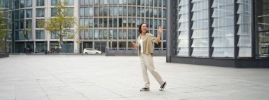 Mutlu Asyalı kız birine veda ediyor, arkadaşına el sallıyor ve akıllı telefonuyla sokakta yürüyor..