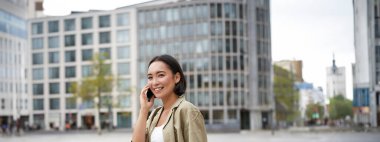 Modern Asyalı genç kız cep telefonuyla konuşuyor, şehir caddesinde telefon kullanıyor. Akıllı telefondan birini ararken gülümseyen bir kadın..