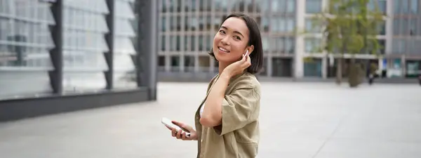 现代人 年轻女人走在城市的大街上 用无线耳机听音乐 拿着智能手机 — 图库照片