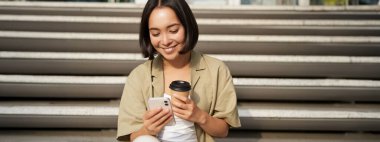 Mutlu şehir kızı hazır kahvesini içiyor ve akıllı telefondan besleniyor. Asyalı kadın elinde cep telefonu, elinde çay ile merdivenlerde oturuyor..
