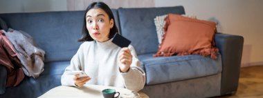 Oturma odasında genç bir kadın, kredi kartını gösteriyor ve ödemek için akıllı telefonu kullanıyor, internetten sipariş veriyor ya da başvuru üzerinden alışveriş yapıyor..