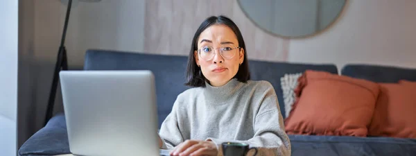 Korean woman with perplexed face, sitting with laptop, working on remote, freelancer with confused expression looking at camera.