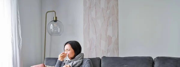 Woman with temperature, coughing, showing symptoms of flu, influenza or cold, sitting in warm clothes on sofa, covered with blanket, sneezing, staying at home in living room.