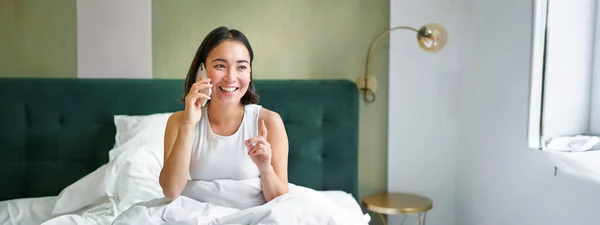 Beautiful smiling asian girl talking on mobile phone, lying in bed with pleased happy face, speaking to someone on telephone.