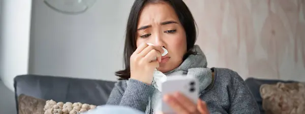 Ill korean woman sitting at home, sneezing, caught cold, reading news on mobile phone, freezing at her home because of heating problems.