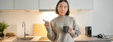 Gülümseyen Asyalı kızın portresi mutfakta duruyor, fincandan kahve içiyor ve afişi işaret ediyor, ajansların reklamını gösteriyor..