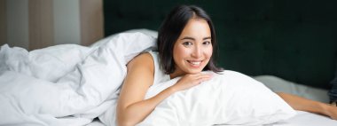 Genç Koreli kadının portresi gülümseyen, yastığa uzanmış, yatak odasında poz veren, rahatlamış ve rahat hisseden.
