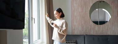 - Günaydın. Mutlu Asyalı kadın evde espresso içiyor, pencereden dışarı bakıyor ve gülümsüyor, sıcak oturma odasında kendini sıcak hissediyor..