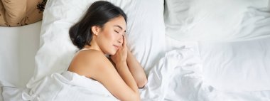 Asyalı bir kadının beyaz yastıklarda kral yatağında tek başına uyuduğu en iyi görüntü. Genç kız yatak odasında gözleri kapalı yatıyor, sabah güneşi odada parlıyor..