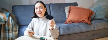 Oturma odasında genç bir kadın, kredi kartını gösteriyor ve ödemek için akıllı telefonu kullanıyor, internetten sipariş veriyor ya da başvuru üzerinden alışveriş yapıyor..