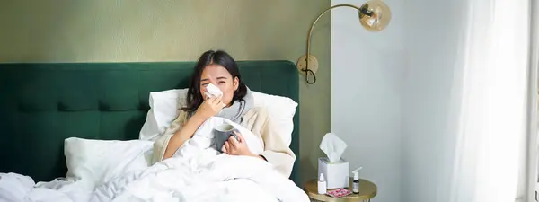 Covid-19, flu and vaccination concept. Korean girl lying in bed with cold, catching influenza, drinking hot tea and using prescribed medication.