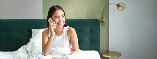Smiling korean girl in bed, talks on mobile phone, making a phone call, lazy morning as asian woman orders delivery via smartphone.