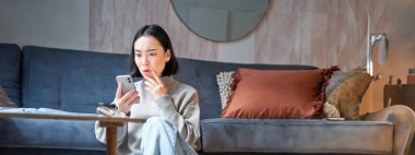 Genç Asyalı kızın dikey görüntüsü cep telefonuna şaşırmış görünüyor, harika haberler okuyor, akıllı telefon uygulamasında bilgilendirme yapıyor, evinde yerde oturuyor..