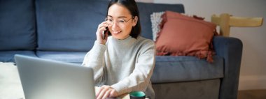 Gülümseyen Asyalı kadının portresi telefon görüşmesi yapıyor, dizüstü bilgisayarda çalışıyor ve akıllı telefondan sohbet ediyor..