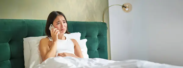 Portrait of concerned asian woman holding mobile phone, receive bad phone call, looking worried and upset, having difficult telephone conversation while lying in bed.