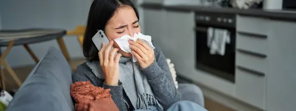 Portrait of ill young korean woman feeling sick, sneezing and holding napkin, staying at home ill, caught cold. Talking on mobile phone.