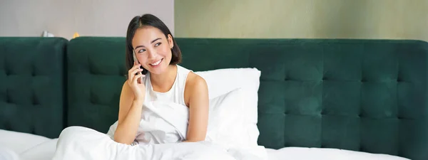 Woman talks on mobile phone in bed. Smiling girl having telephone conversation while relaxing in bedroom.