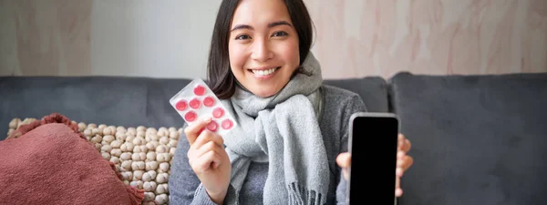 Smiling korean girl shows smartphone screen, medication in hands, feeling sick and staying at home, using online GP doctor app while on sick leave.