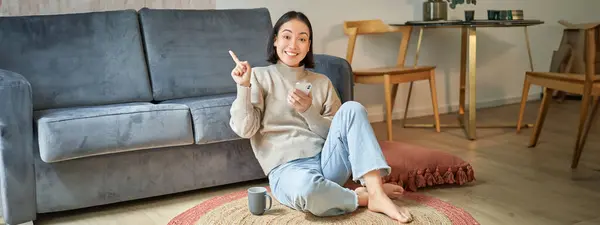 Smiling asian girl sits on floor in stylish living room, pointing finger at advertisement, showing promo banner, holding mobile phone in hand.