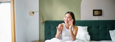 Güzel gülümseyen Asyalı kız cep telefonuyla konuşuyor, mutlu bir yüzle yatakta uzanıyor, telefonda biriyle konuşuyor..