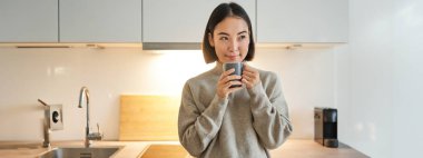 Gülümseyen Asyalı kadının portresi mutfağında duruyor, kahve içiyor ve kameraya bakıyor, rahat ve rahat ev kavramı..