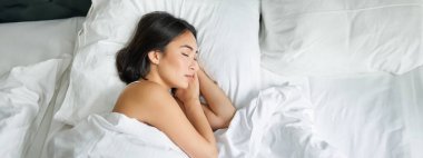 Asyalı bir kadının beyaz yastıklarda kral yatağında tek başına uyuduğu en iyi görüntü. Genç kız yatak odasında gözleri kapalı yatıyor, sabah güneşi odada parlıyor..