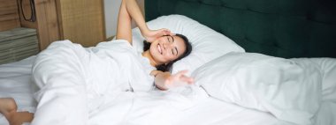 Romantik Asyalı kız rahat bir yatak odasında uyanıyor, beyaz çarşaflarla yatakta uzanıyor, elini boş yastığa uzatıyor..