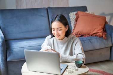 Evden çalışmak, serbest çalışmak ve e-öğrenme konsepti. Çalışan genç bir kadın, dizüstü bilgisayarın önünde oturuyor, uzaktan kumandayla çalışıyor, oturma odasında çalışıyor..