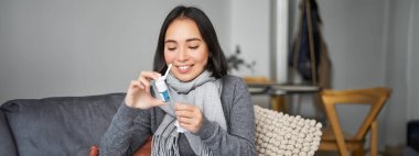 Boğazı ağrıyan bir kadın spreyine bakıyor, soğuk algınlığı tedavisi görüyor, grip sonrası evde kalıyor, kendini daha iyi hissediyor, oturma odasında eşarpta oturuyor..