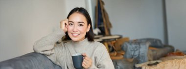 Gülümseyen Asyalı kadın elinde kupasıyla kanepede oturuyor, evde kahve içiyor ve işten sonra dinleniyor, sakin ve rahat görünüyor..