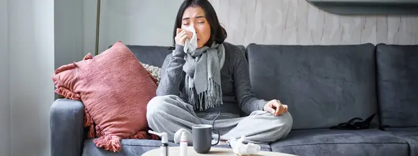 Portrait of korean woman sneezing, feeling sick, staying at home with flu or cold, neck wrapped with scarf, staying at home, taking medication.